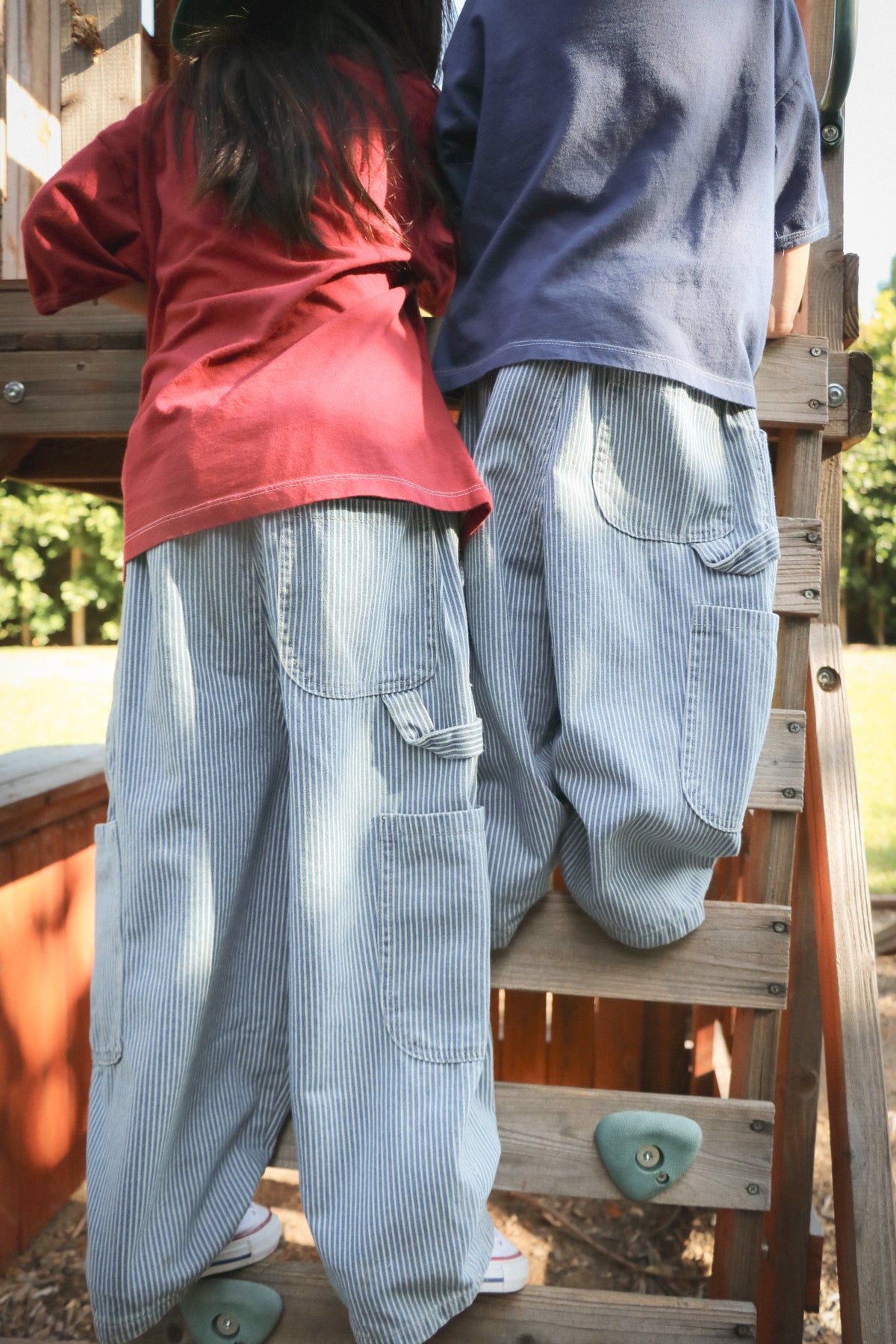 Two children wearing baggy hickory striped jeans climbing ladder up to treehouse